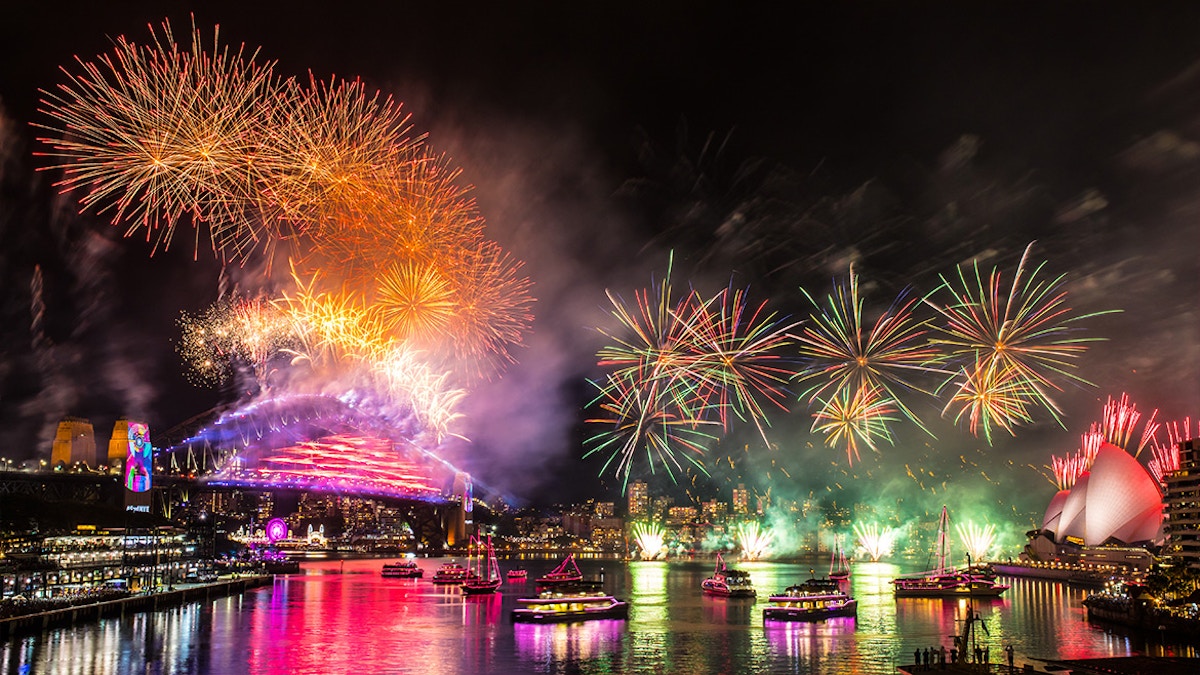 Fireworks light up the night sky over Sydney Harbour, with the Sydney Opera House and Harbour Bridge visible. Boats are on the water and bright reflections color the surface.