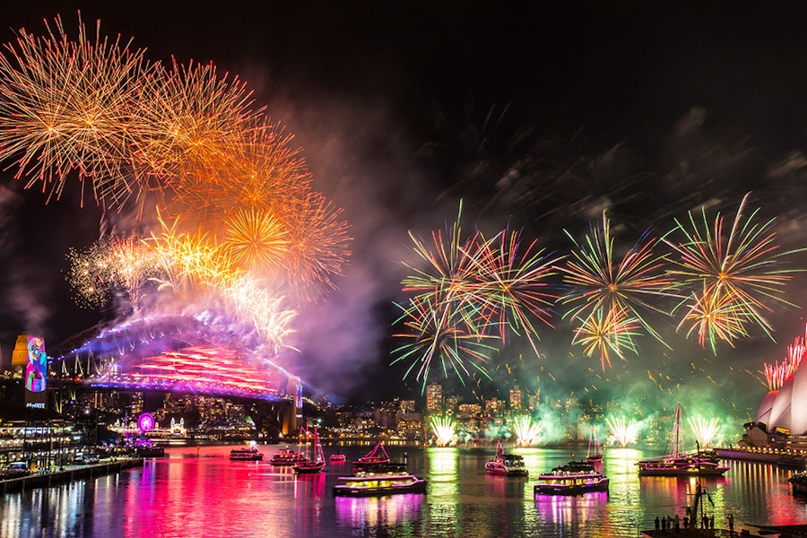 Fireworks light up the night sky over Sydney Harbour, with the Sydney Opera House and Harbour Bridge visible. Boats are on the water and bright reflections color the surface.