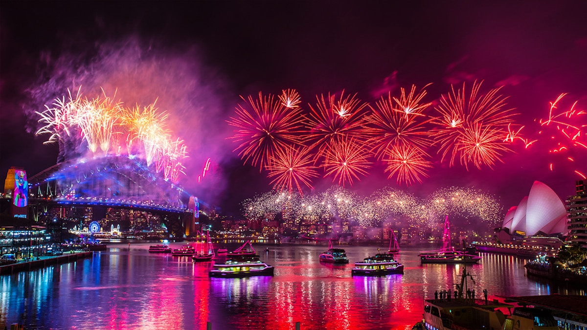 Fireworks light up the night sky above Sydney Harbour Bridge and Sydney Opera House, reflecting on the water with boats below.