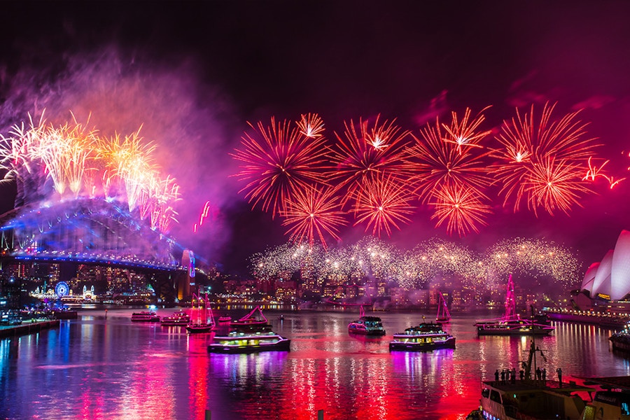 Fireworks light up the night sky above Sydney Harbour Bridge and Sydney Opera House, reflecting on the water with boats below.