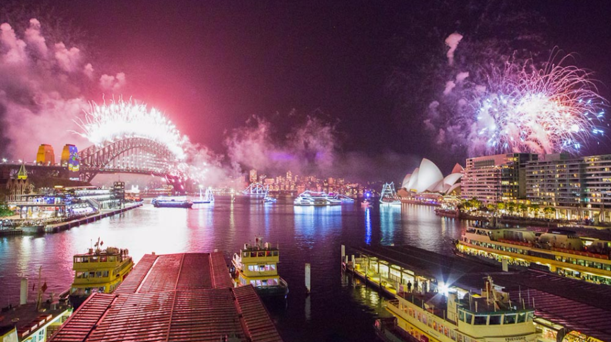Sydney Harbour at night with fireworks over the Harbour Bridge and Opera House, ferries docked in the foreground, and city lights in the distance.