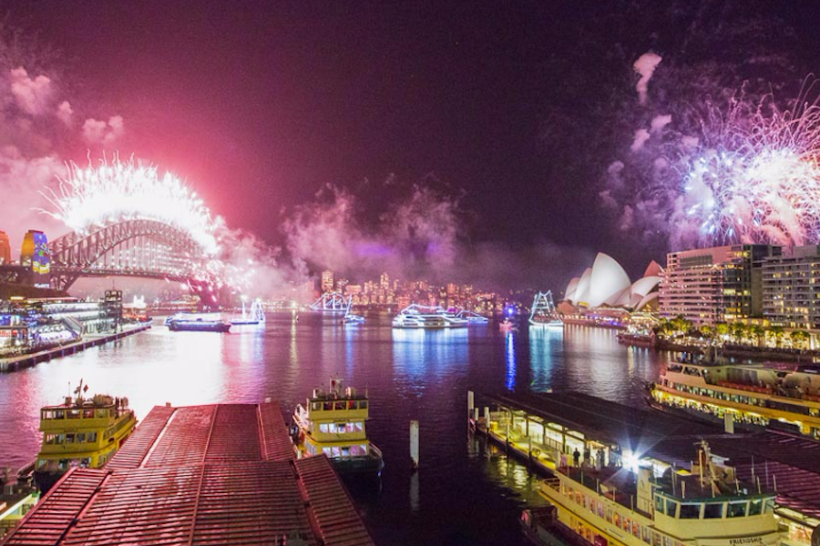 Sydney Harbour at night with fireworks over the Harbour Bridge and Opera House, ferries docked in the foreground, and city lights in the distance.