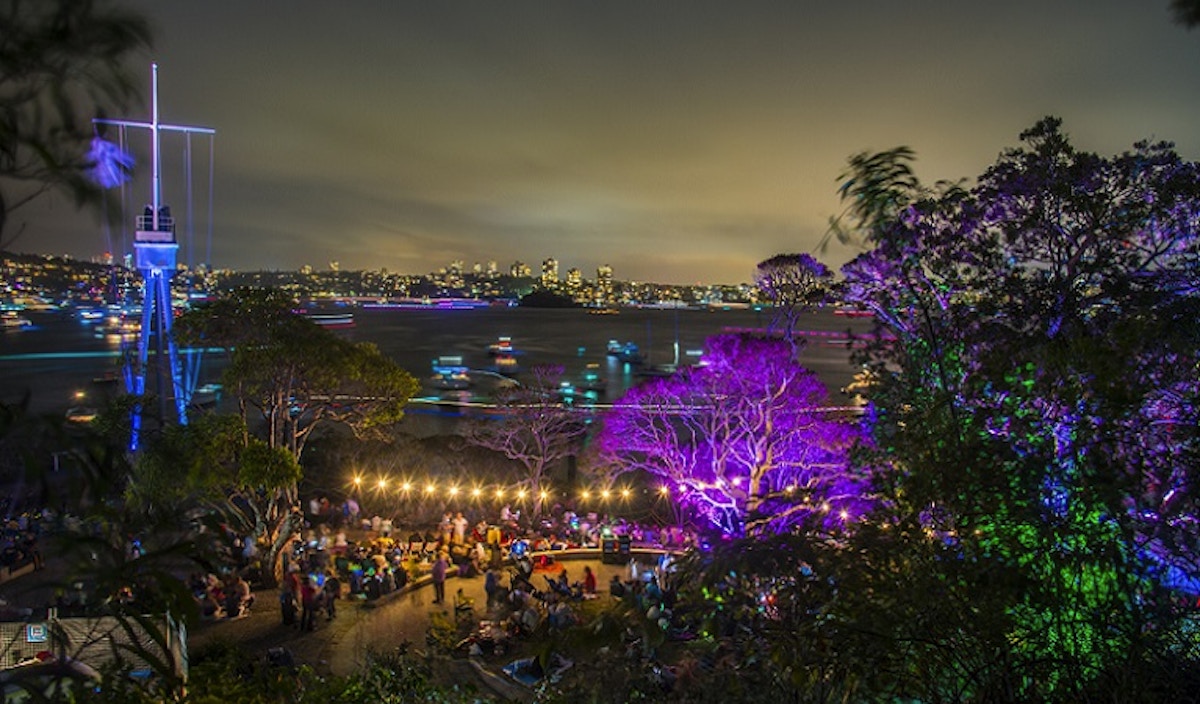 Outdoor night scene with colorful lights illuminating trees and people gathered at an event; city skyline and boats visible on the water in the background.