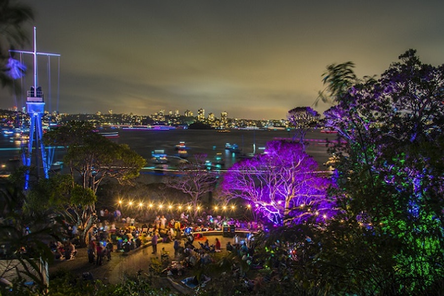 Outdoor night scene with colorful lights illuminating trees and people gathered at an event; city skyline and boats visible on the water in the background.