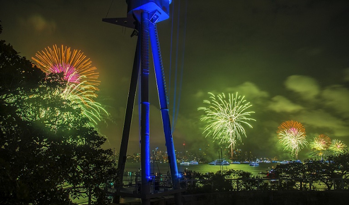 Colorful fireworks light up the night sky over a body of water, with boats visible and silhouetted structures and trees in the foreground.