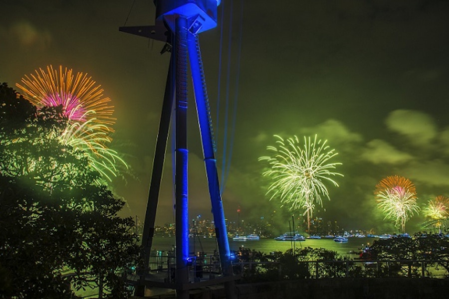 Colorful fireworks light up the night sky over a body of water, with boats visible and silhouetted structures and trees in the foreground.