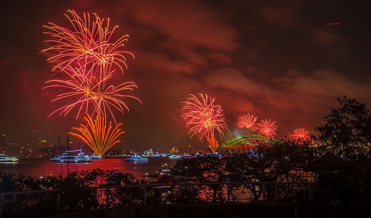 Red fireworks explode over a harbor at night, with boats on the water and a bridge visible in the background.