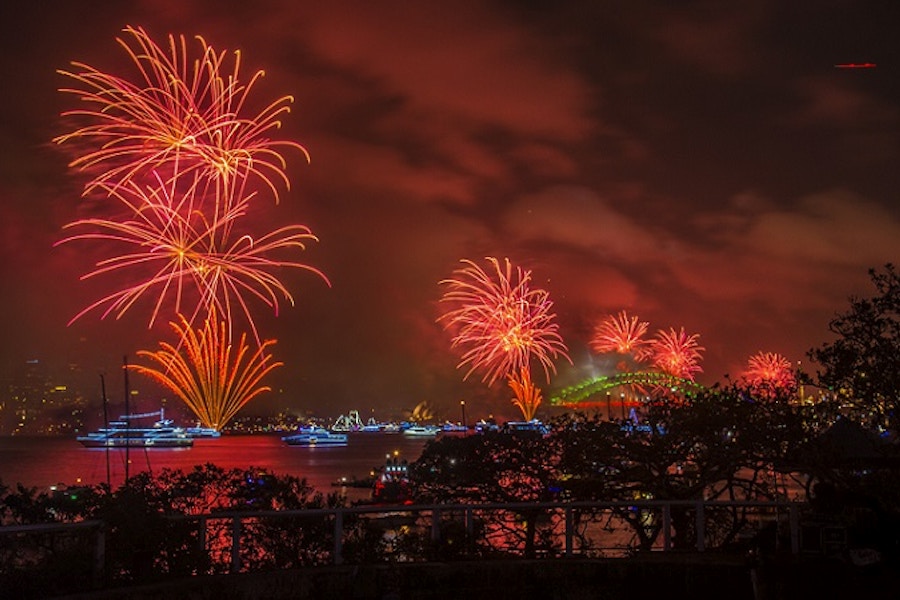 Red fireworks explode over a harbor at night, with boats on the water and a bridge visible in the background.