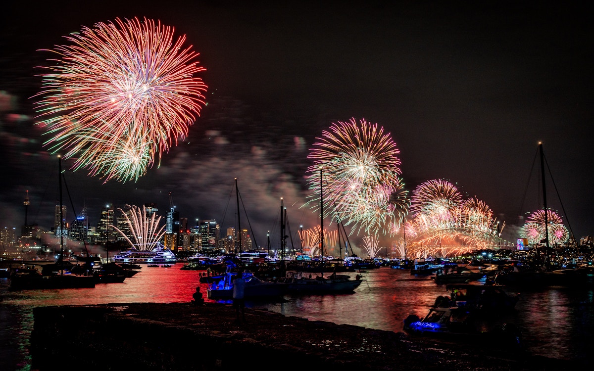 Fireworks light up the night sky above a city skyline, reflecting on water with boats gathered below.