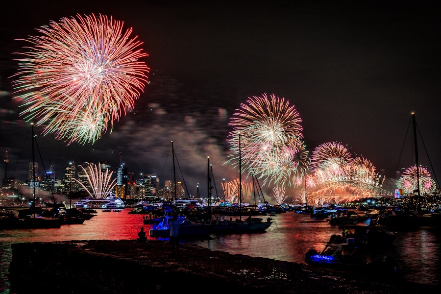 Fireworks light up the night sky above a city skyline, reflecting on water with boats gathered below.
