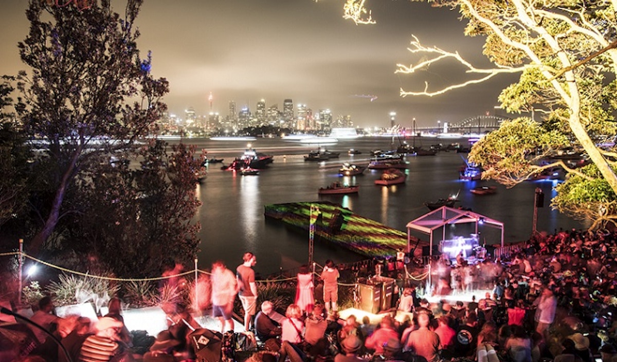 A crowd gathers by the waterfront at night, watching boats on the water with a city skyline illuminated in the background.