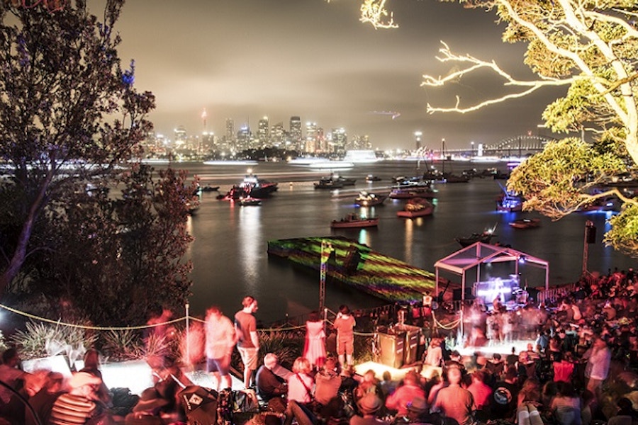A crowd gathers by the waterfront at night, watching boats on the water with a city skyline illuminated in the background.