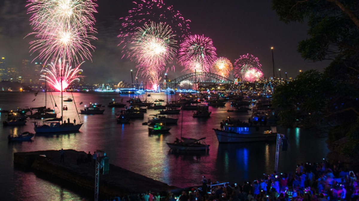 Fireworks light up the night sky above a harbor filled with boats, with a bridge in the background and a crowd gathered along the water’s edge.