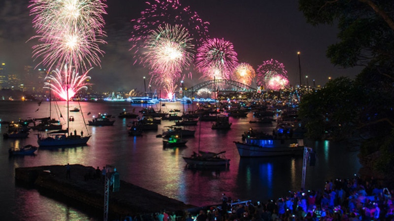 Fireworks light up the night sky above a harbor filled with boats, with a bridge in the background and a crowd gathered along the water’s edge.