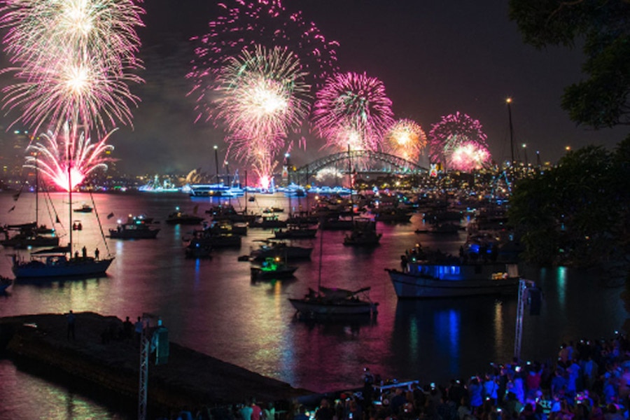 Fireworks light up the night sky above a harbor filled with boats, with a bridge in the background and a crowd gathered along the water’s edge.