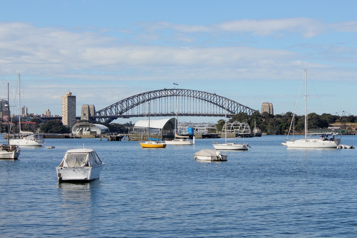 Boats floating on water with the Sydney Harbour Bridge and nearby buildings in the background under a partly cloudy sky.