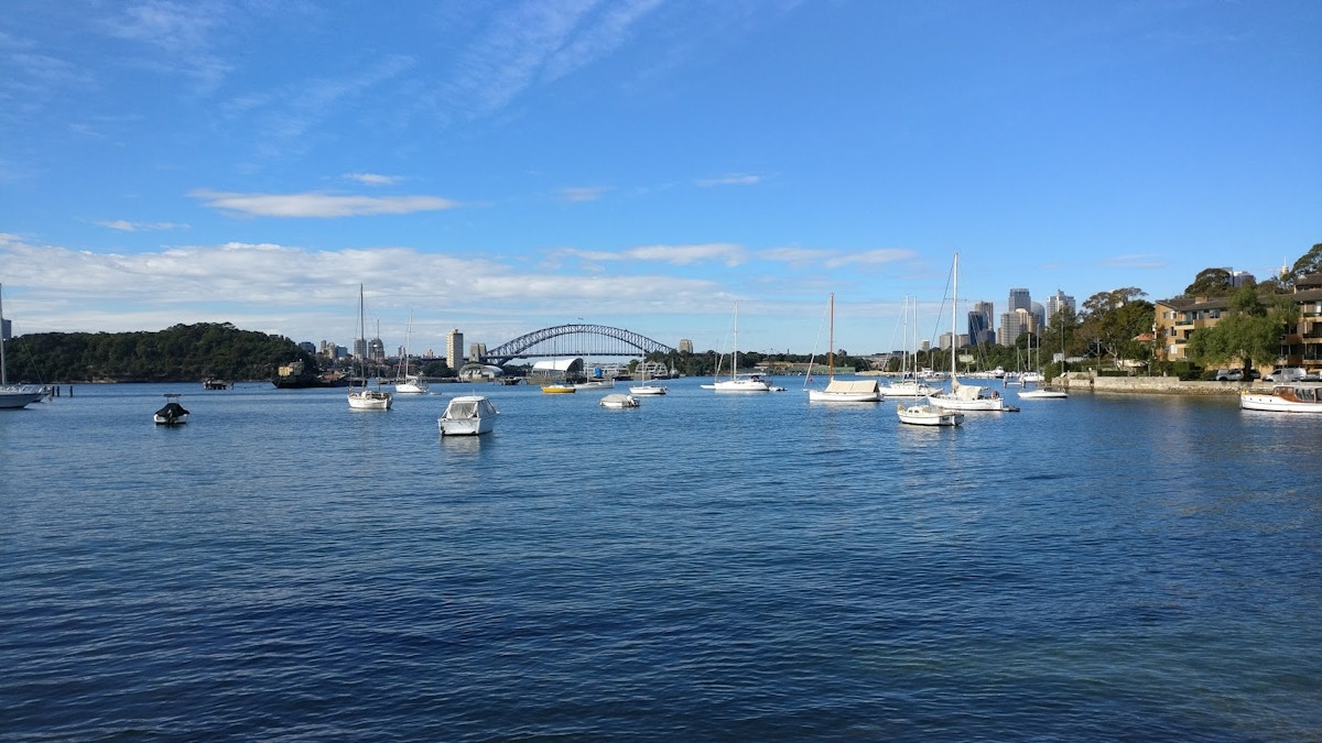 Boats anchored on calm water with Sydney Harbour Bridge and city buildings visible in the background under a clear blue sky.