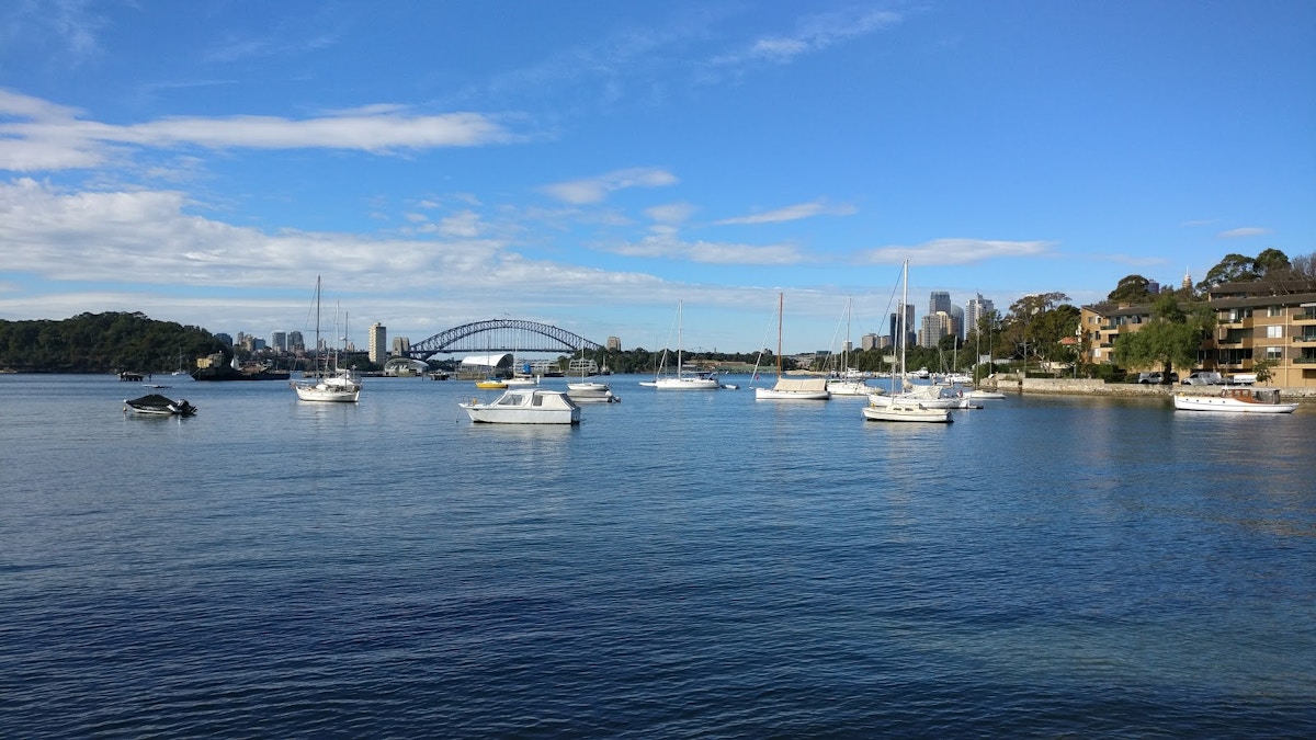 Several boats float on calm water with the Sydney Harbour Bridge and city buildings visible in the background under a blue sky.