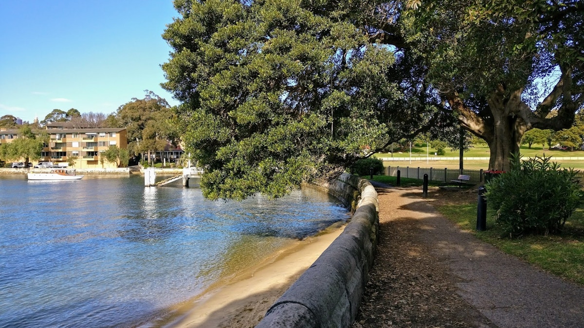 A paved path runs beside a stone wall along a calm waterfront, with large trees, a grassy park, and buildings visible in the background.