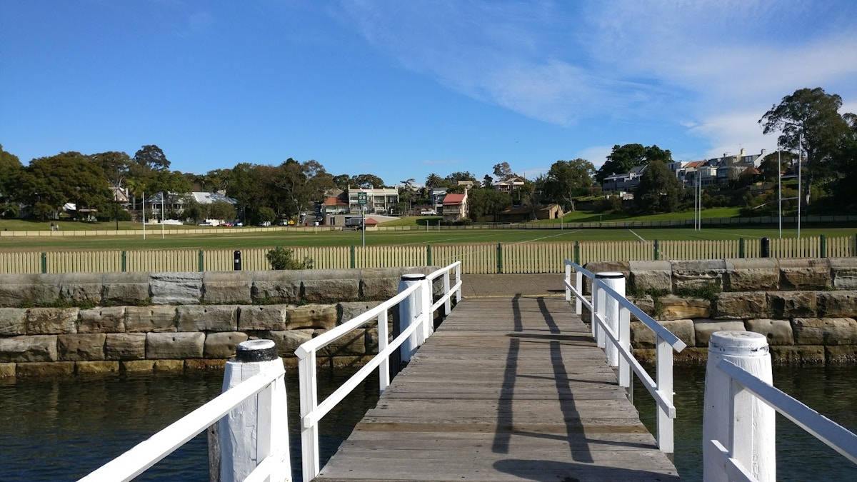 A wooden pier with white railings leads to a grassy park area, bordered by a stone wall and fence, under a clear blue sky.