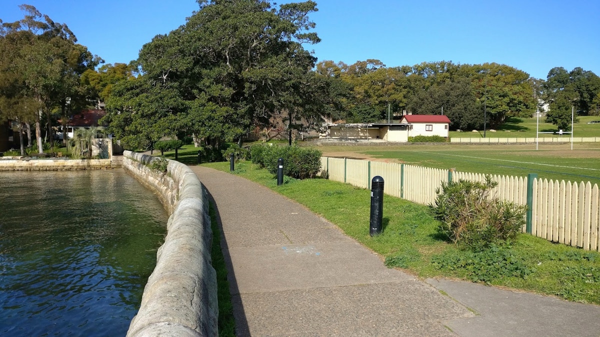 Paved walkway beside a stone wall borders a body of water, with a grassy field, trees, and small buildings in the background under a clear blue sky.