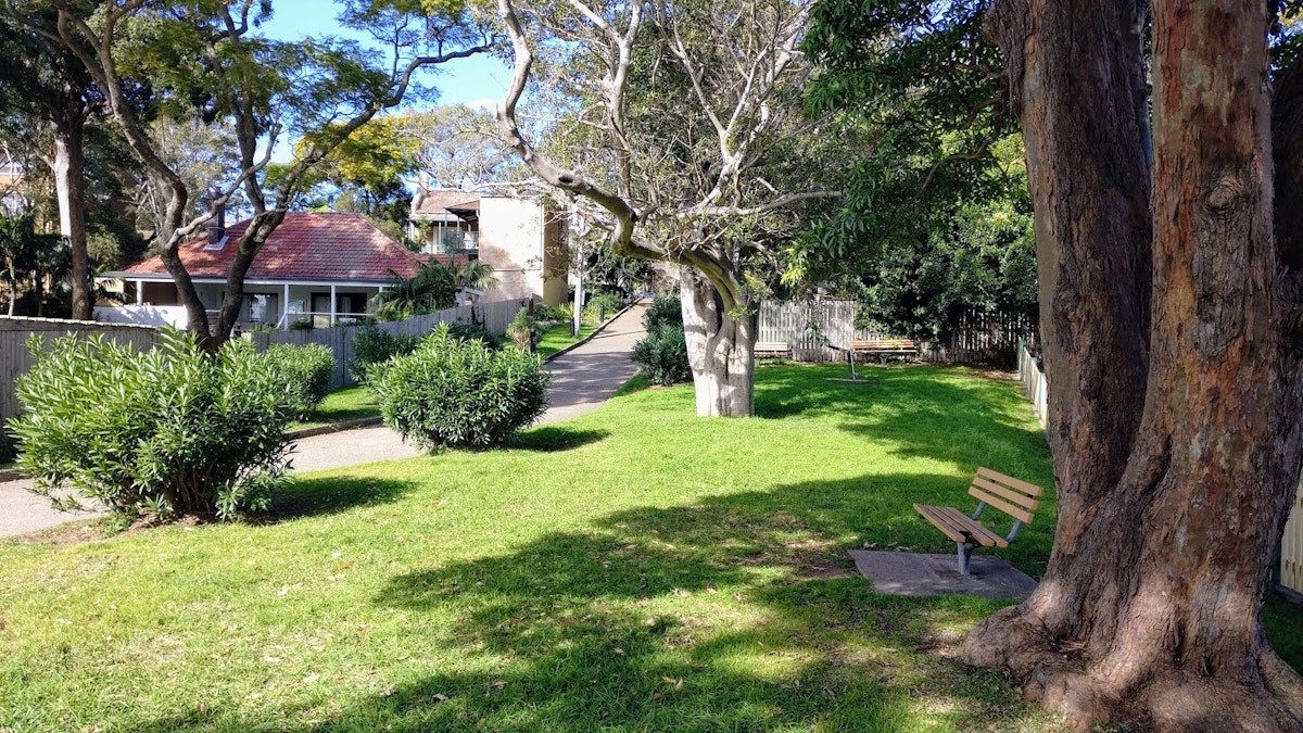 A park with green grass, trees, two benches, a paved path, and houses with red roofs in the background under a clear sky.
