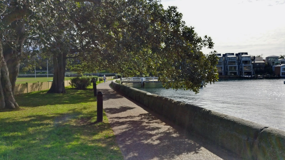 Paved path beside a stone wall and waterway, shaded by a large tree, with grass, buildings, and a footbridge in the background.