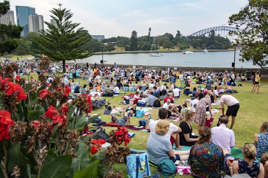 A large crowd sits on blankets in a grassy park near the water, with trees, boats, and a bridge visible in the background on a partly cloudy day.