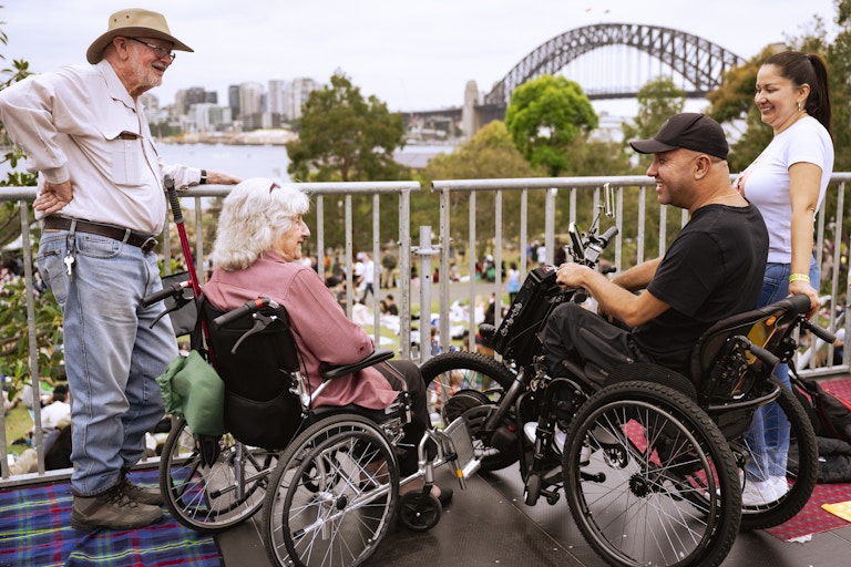 Two people in wheelchairs and two standing individuals converse on a platform overlooking a cityscape and a large bridge, with a crowd below.