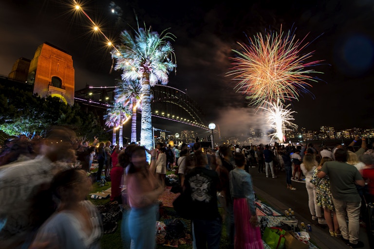 A crowd watches colourful fireworks light up the night sky near Sydney Harbour Bridge, with palm trees and city lights visible.
