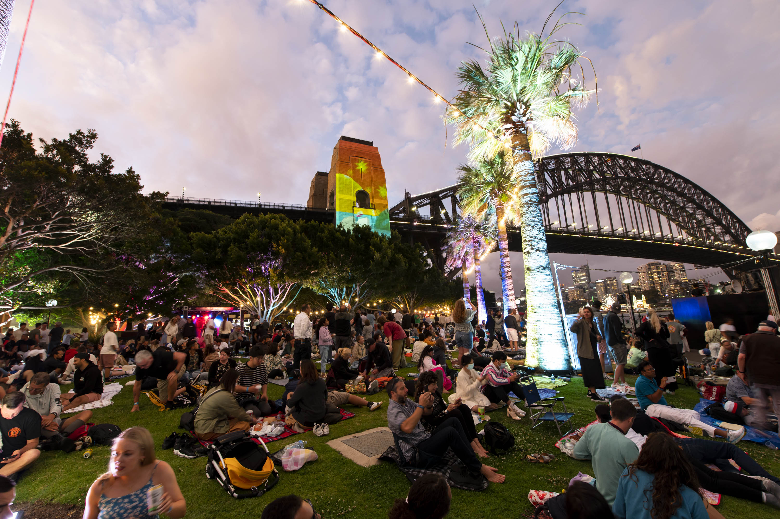 A large crowd sits on grass at dusk near the Sydney Harbour Bridge, with trees and a palm tree illuminated by colourful lights.