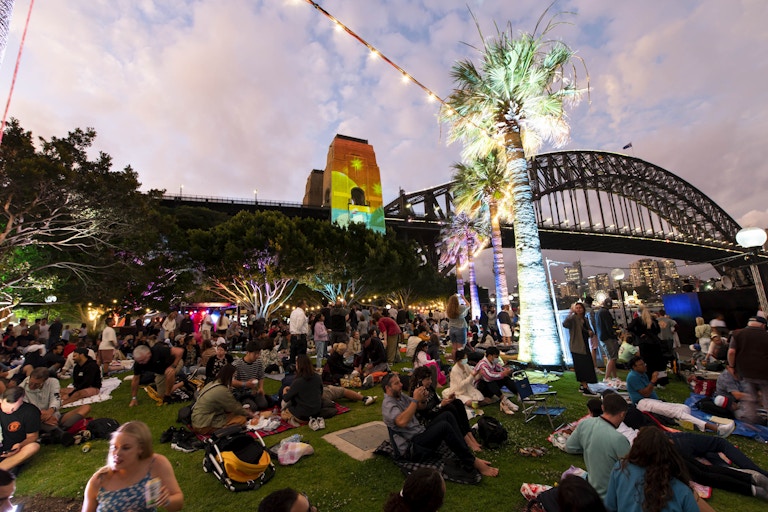 A large crowd sits on grass at dusk near the Sydney Harbour Bridge, with trees and a palm tree illuminated by colourful lights.