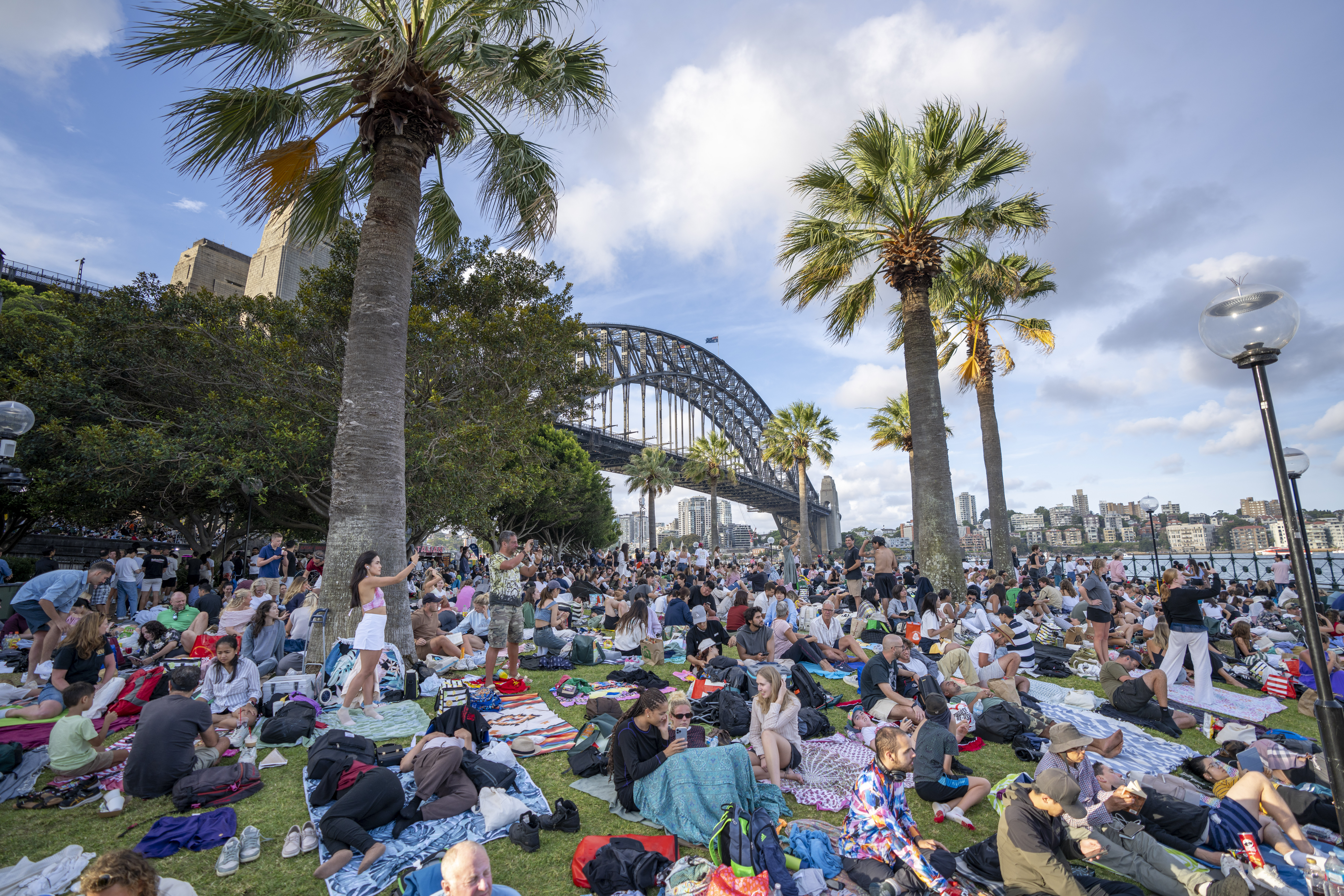 A large group of people sitting on the grass in front of a bridge.
