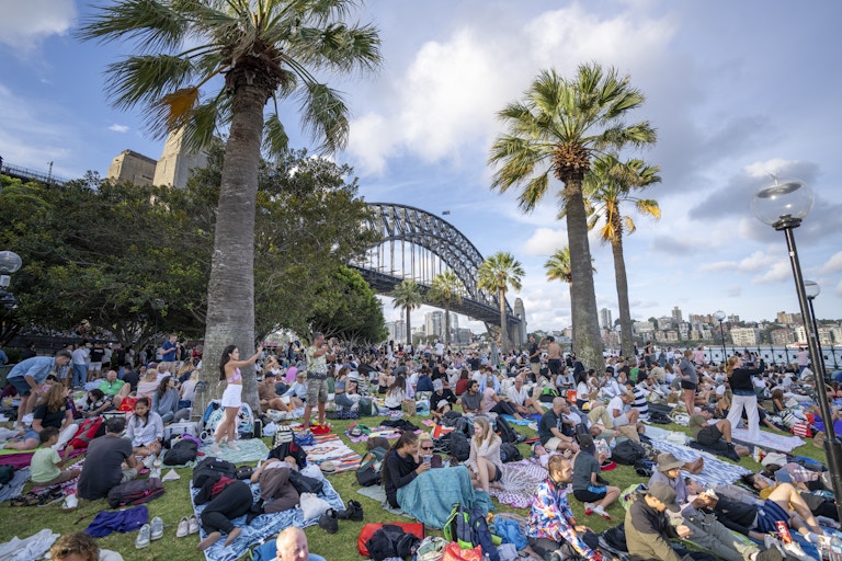 A large group of people sitting on the grass in front of a bridge.