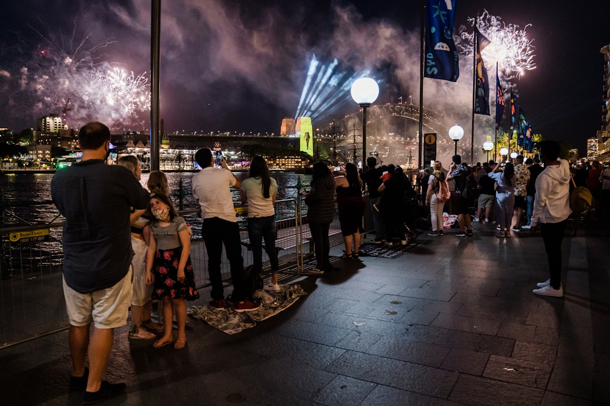 People watch fireworks over a city harbor at night; some stand by a railing, others sit on blankets. Bright lights and smoke fill the sky, with city buildings and a lit bridge in the background.