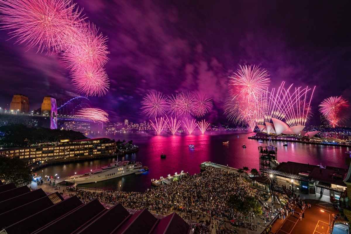 A large crowd watches vibrant pink fireworks explode over Sydney Harbour, with the Sydney Opera House and Harbour Bridge illuminated at night.