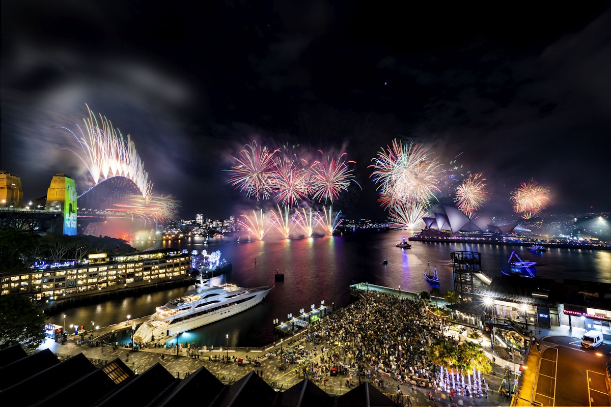 Fireworks light up the night sky over Sydney Harbour, with crowds gathered near the Opera House and Harbour Bridge, and boats docked in the water.