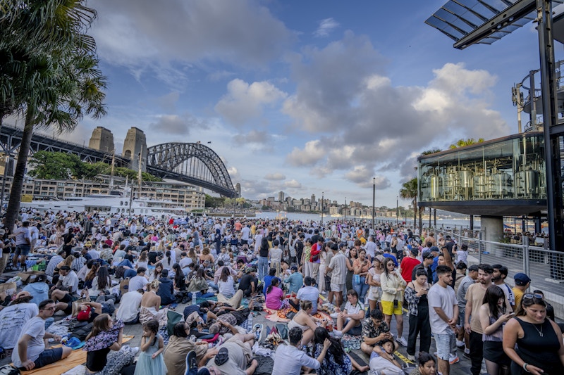 Large crowd gathers outdoors near Sydney Harbour Bridge during the day, with people sitting and standing under partly cloudy skies.