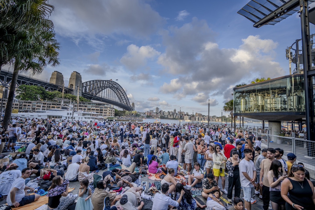 Large crowd gathers outdoors near Sydney Harbour Bridge during the day, with people sitting and standing under partly cloudy skies.