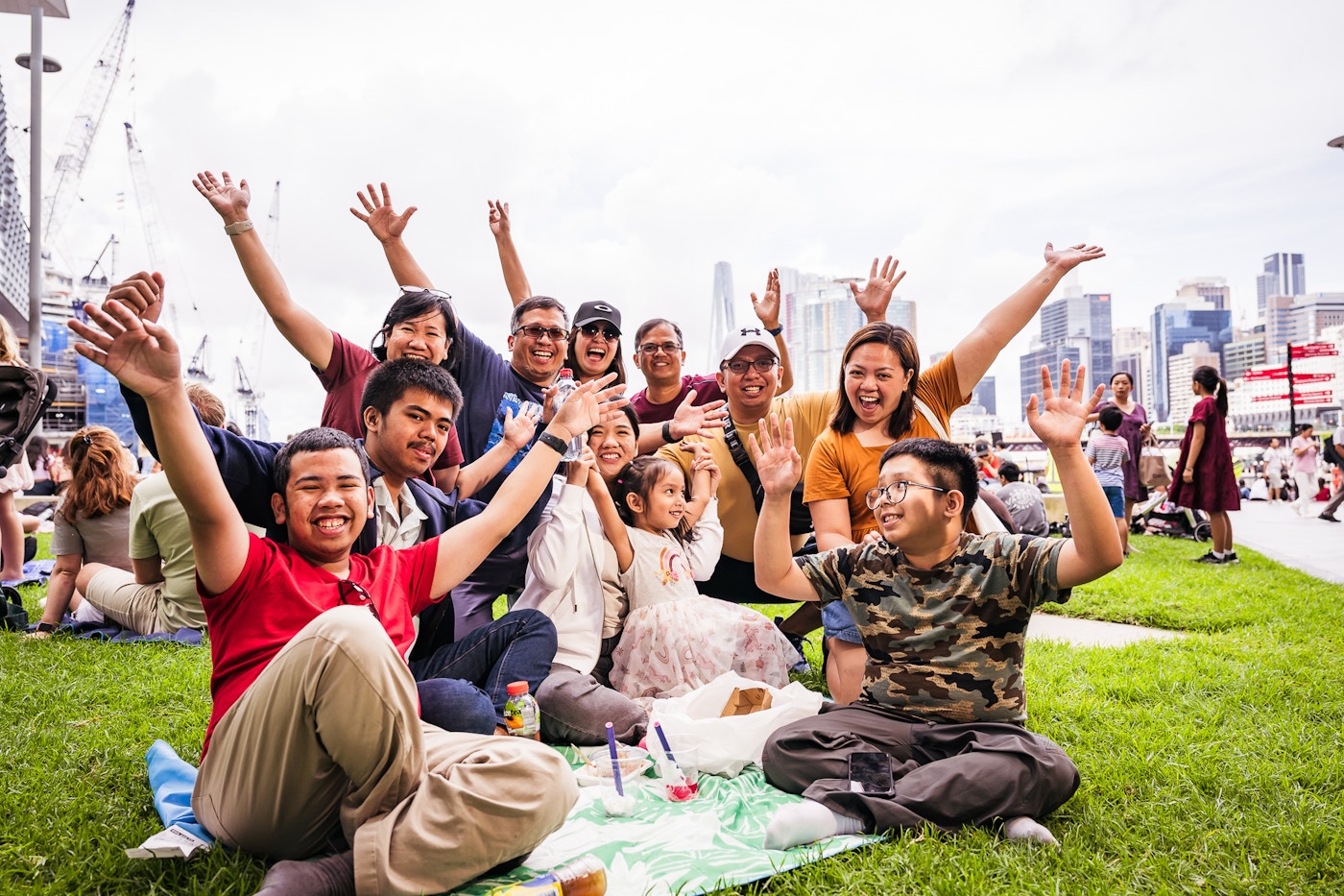 A group of people sitting on grass outdoors smile and raise their arms, with a city skyline and other people in the background.