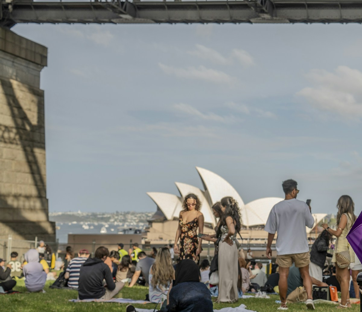 People sit and stand on grass beneath a bridge, with the Sydney Opera House visible in the background under a partly cloudy sky.