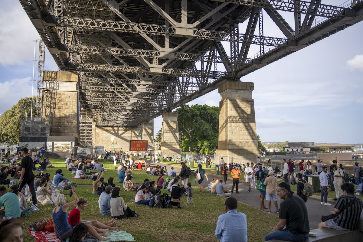 People gather and relax on grass under a large steel bridge, with some sitting on blankets and others walking around on a sunny day.