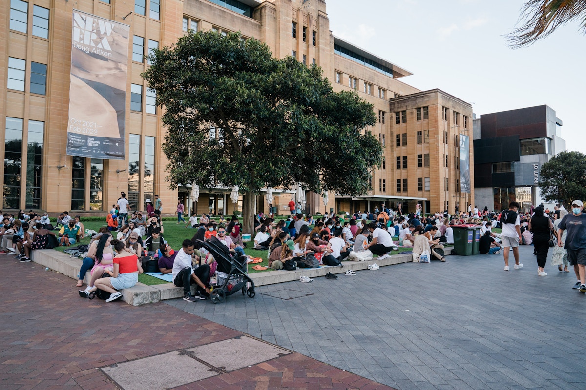 A large group of people sit and gather outside a tan stone building with trees and banners, enjoying a sunny day.