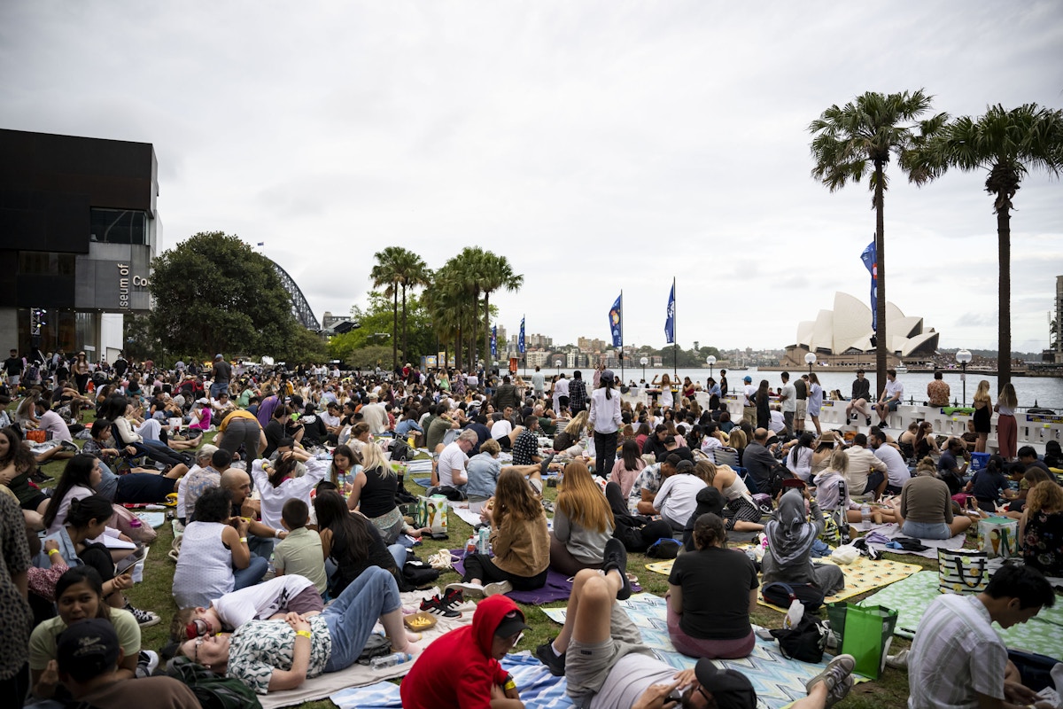 A large crowd sits on blankets in a park near the waterfront, with palm trees and the Sydney Opera House visible in the background under a cloudy sky.