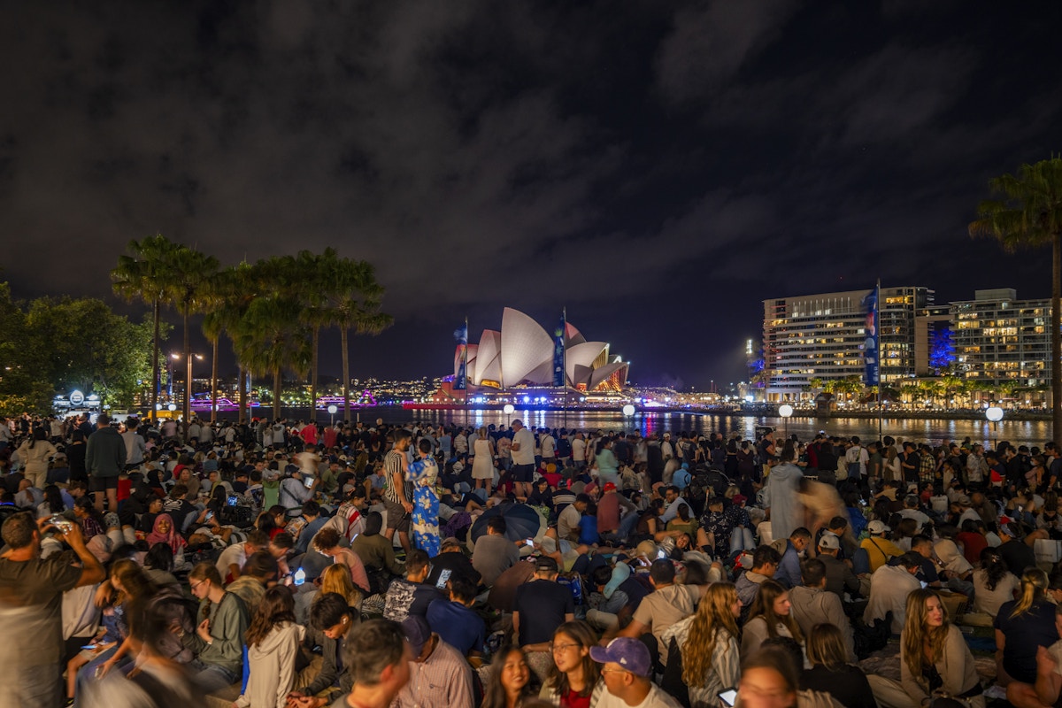 A large crowd sits on the ground at night facing the Sydney Opera House, which is illuminated across the harbor. City buildings and trees are visible in the background.