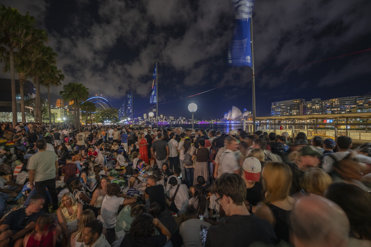 Large crowd gathers at night near Sydney Harbour; the Sydney Opera House and Harbour Bridge are illuminated in the background.