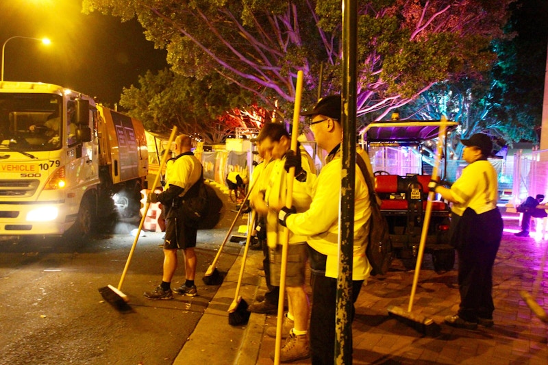A group of workers in yellow vests stand on a street at night, holding brooms, with a truck and colorful tree lights in the background.