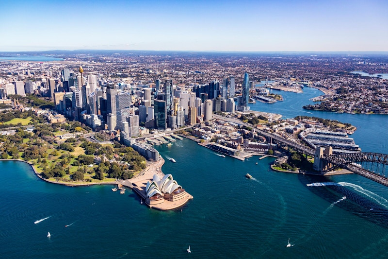 Aerial view of Sydney, featuring the Opera House, skyscrapers, waterways, and surrounding green spaces under a clear blue sky.