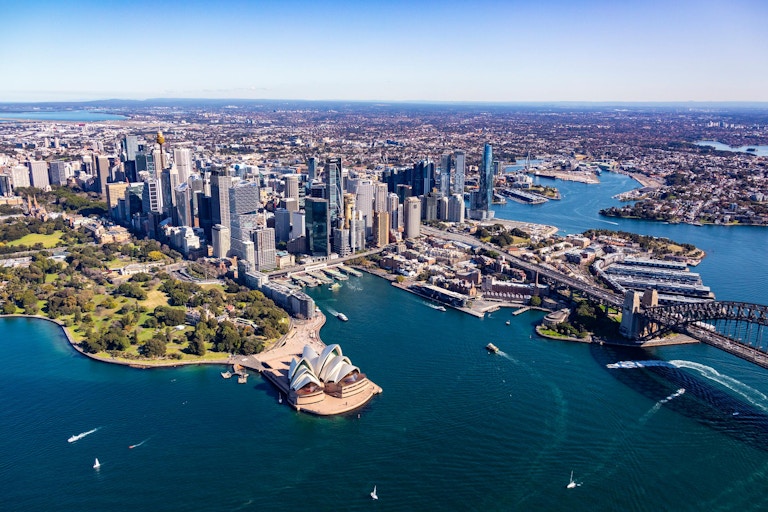 Aerial view of Sydney, featuring the Opera House, skyscrapers, waterways, and surrounding green spaces under a clear blue sky.