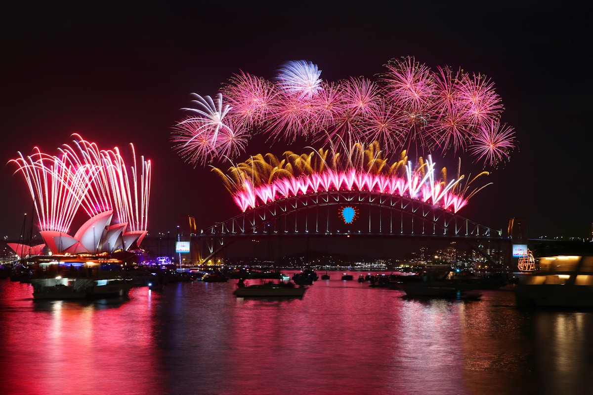 Fireworks illuminate the night sky above the Sydney Opera House and Sydney Harbour Bridge, reflecting on the water with boats in the foreground.